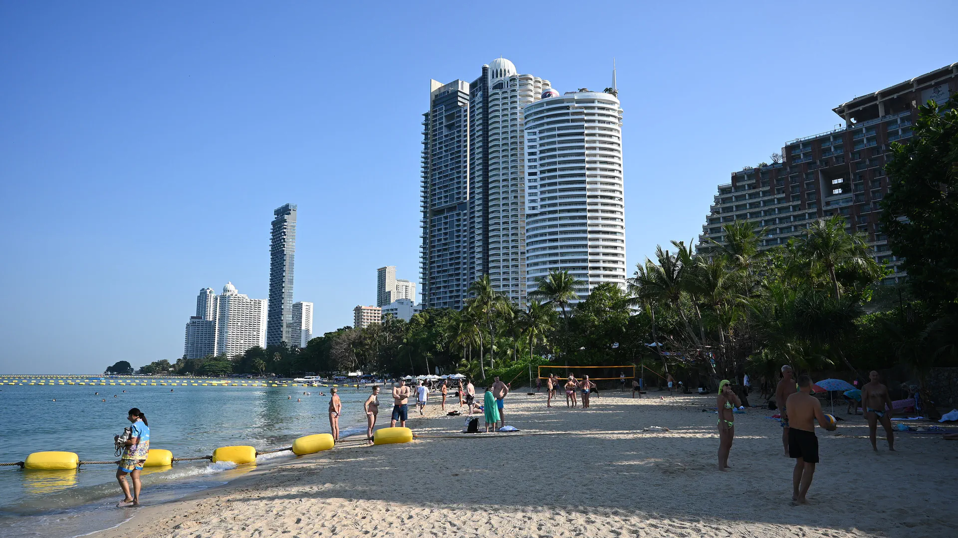 People relax on a sunny Pattaya beach beneath palm trees and high-rise buildings.
